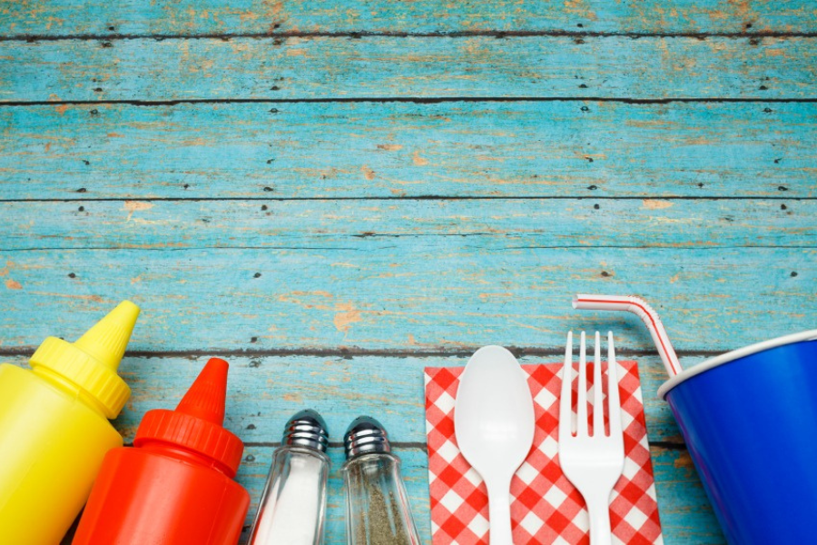 Image of mustard bottle, ketchup bottle, salt and pepper shakers, spoon on red and white checkered napkin, laid out on top of a blue faded wooden picnic table.
