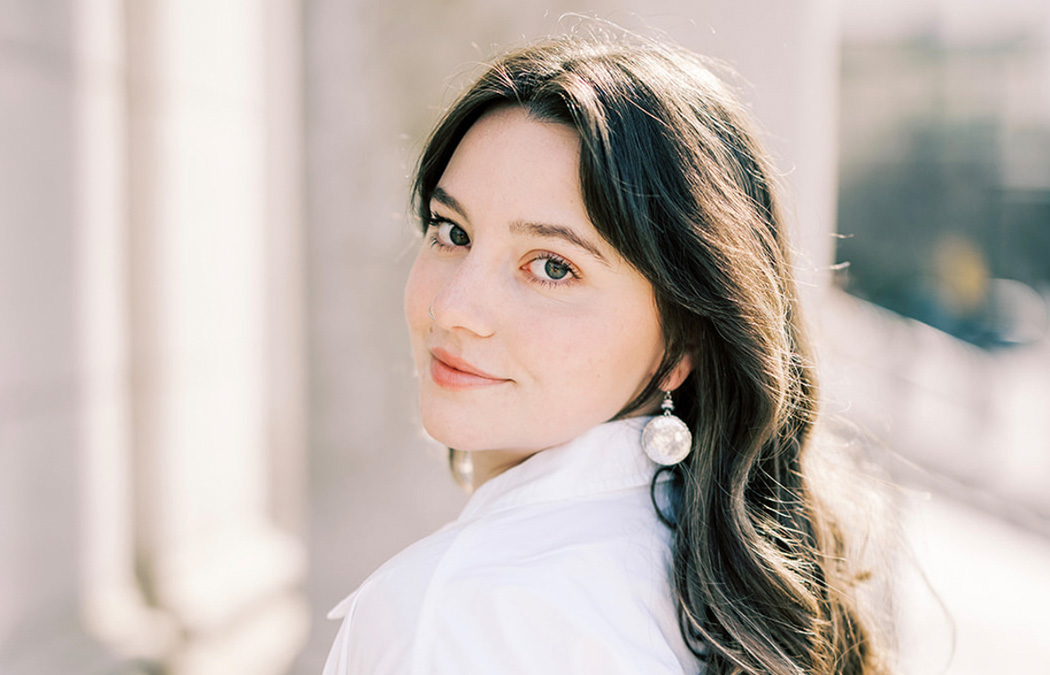 Woman with long brown hair, head turned over shoulder and slightly smiling while looking straight ahead.