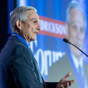 A man turned to his side speaks into a microphone. An image on a projector is visible in the background.
