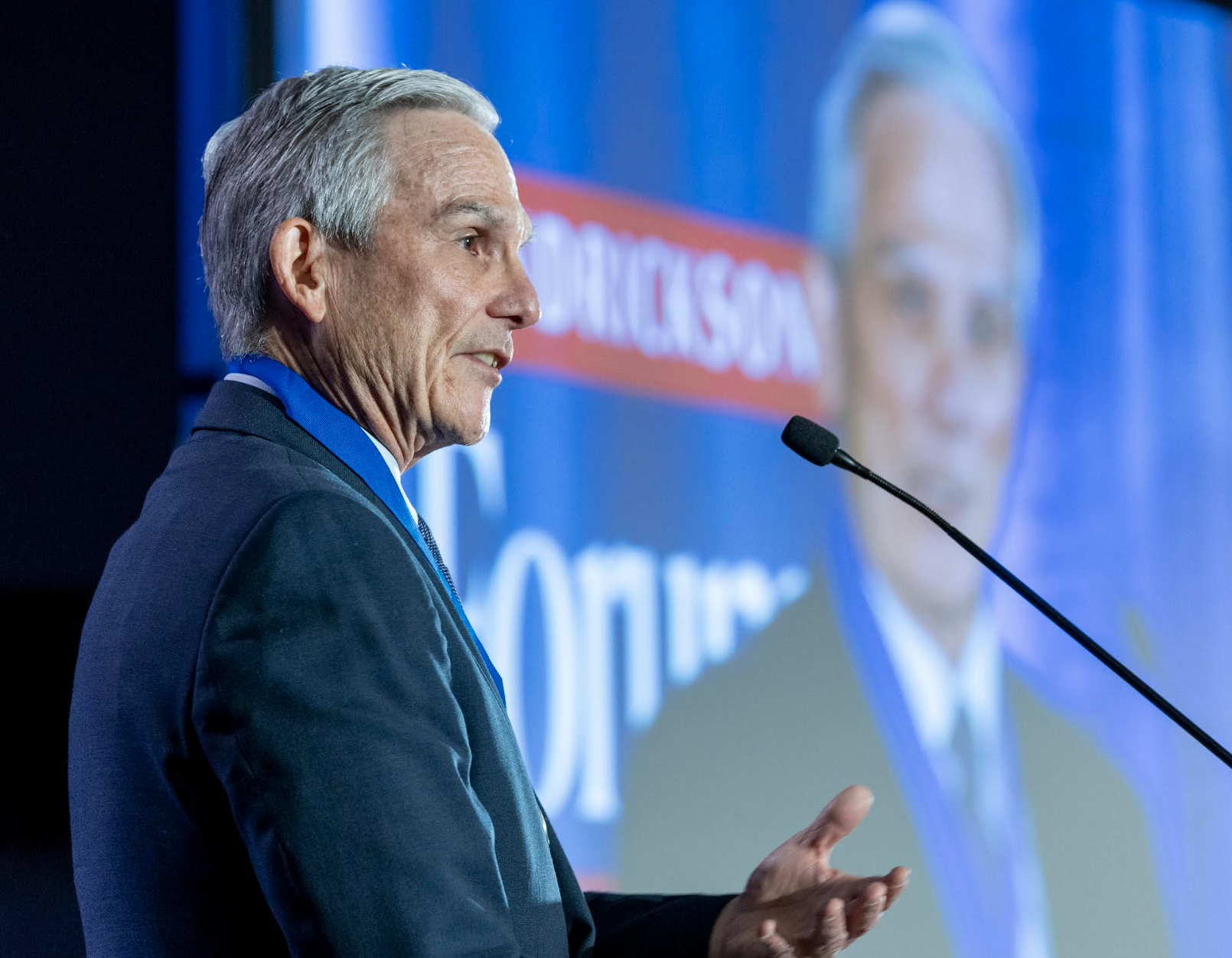 A man turned to his side speaks into a microphone. An image on a projector is visible in the background.