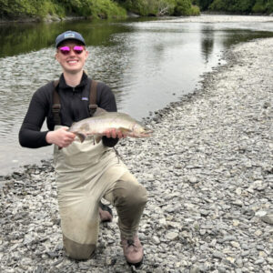 A person kneels on a rocky riverbank, smiling and holding a large fish. They're wearing sunglasses, a cap, and waders. The river flows gently in the background, bordered by lush greenery.