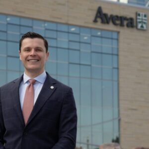 Smiling man in dark suit with white shirt and salmon pink tie, standing out front of a building with glass windows and the text "Avera" at the top, affixed to the building.
