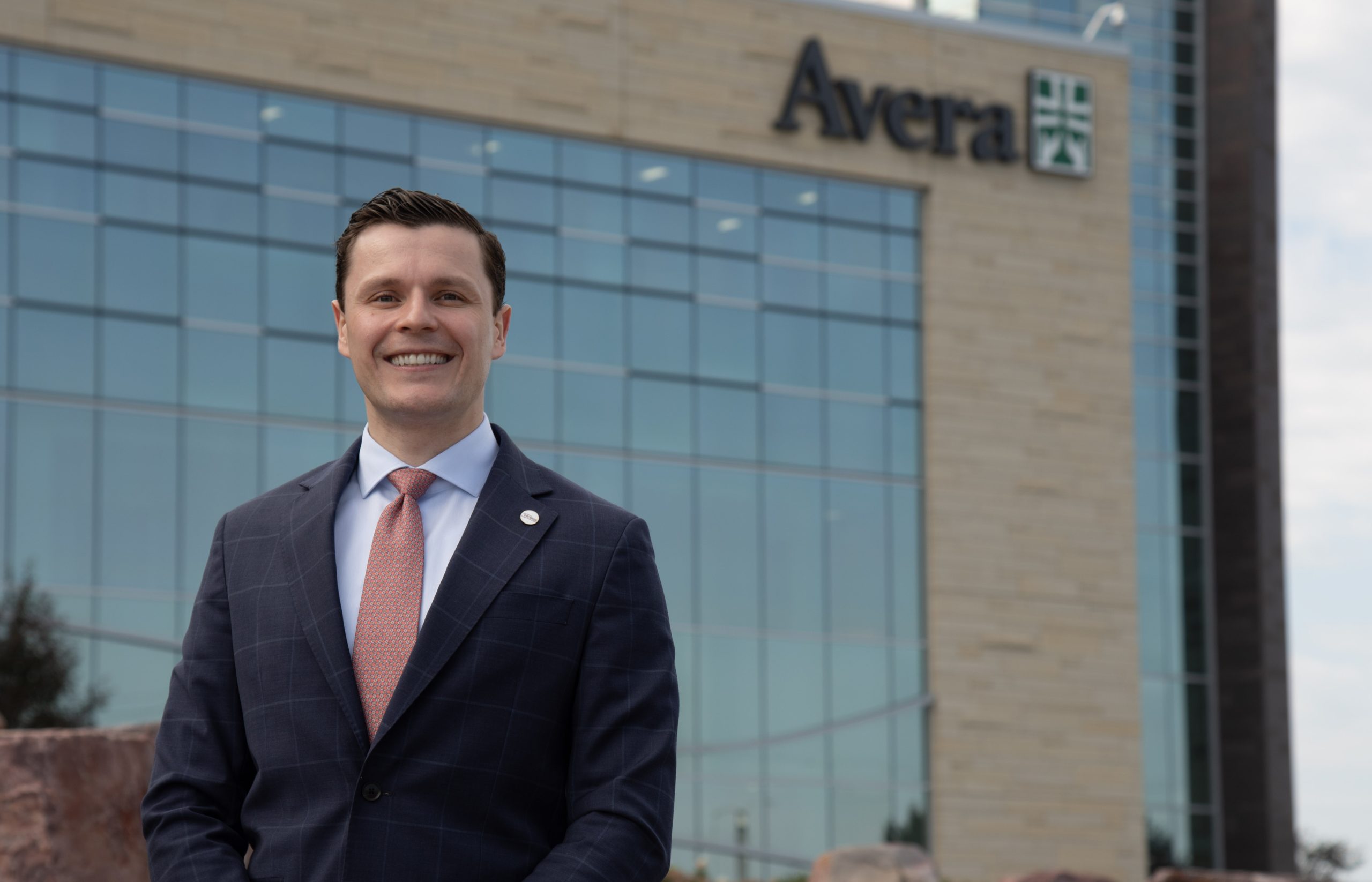 Smiling man in dark suit with white shirt and salmon pink tie, standing out front of a building with glass windows and the text "Avera" at the top, affixed to the building.