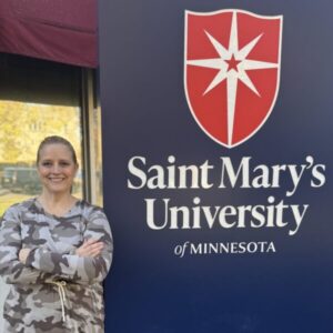 A person in a camo-patterned hoodie stands with arms crossed beside a sign for Saint Mary's University of Minnesota. The sign displays the university's logo and name.