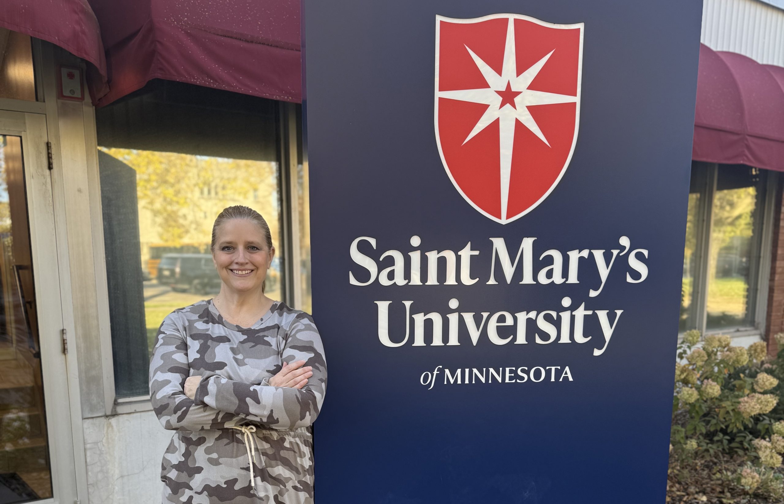 A person in a camo-patterned hoodie stands with arms crossed beside a sign for Saint Mary's University of Minnesota. The sign displays the university's logo and name.