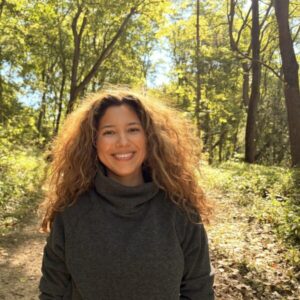 A woman with long, curly hair smiles while standing on a sunlit forest trail.