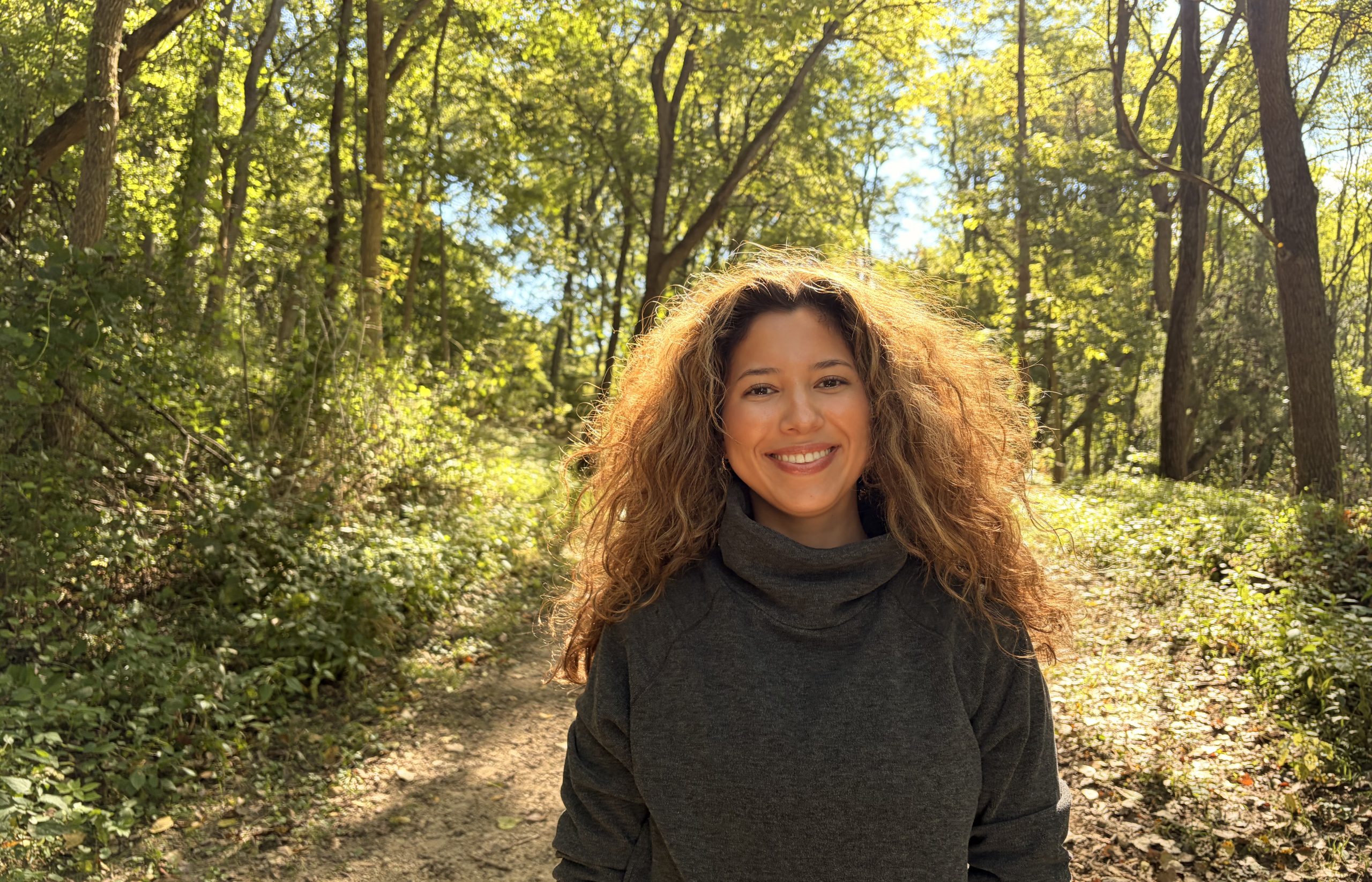 A woman with long, curly hair smiles while standing on a sunlit forest trail.
